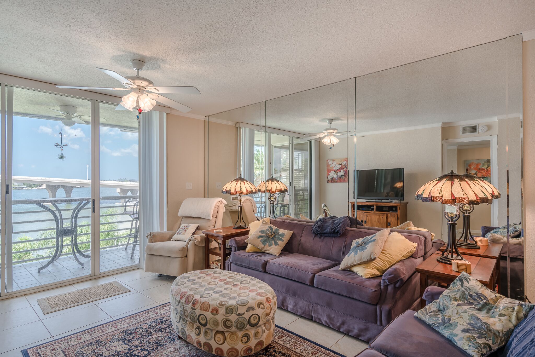A spacious living room with a large sofa, ottoman, and a television. There is a balcony with a view of a bridge and greenery.