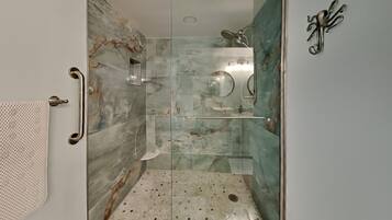 A walk-in shower with a glass door, marble walls, a handheld showerhead, a handheld shower head, a towel rack, and a small shelf.