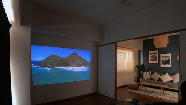 A living room with a large screen displaying a beach scene, a sofa, a coffee table, and a sliding door leading to another room. - OUCHI HOTEL Hijiyama (Hiroshima)