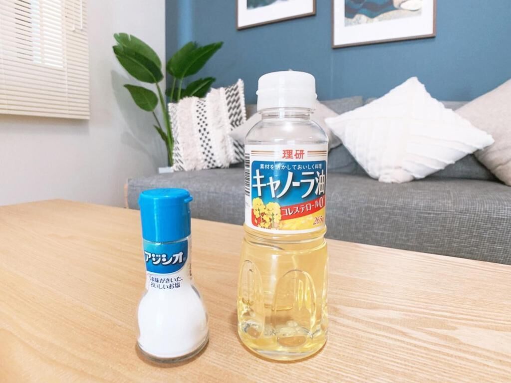 A bottle of liquid and a smaller bottle on a wooden table in a room with a grey sofa and a plant in the background.