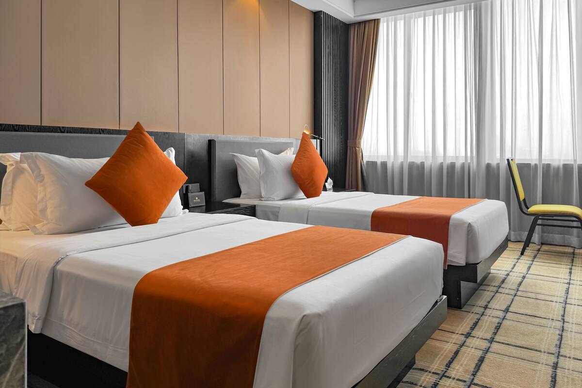 Ein Hotelzimmer mit zwei Betten, jeweils mit weißer Bettwäsche und orangefarbenen Akzenten, einem modernen Kopfteil, einem Nachttisch mit Lampe und Blick auf die Stadt durch transparente Vorhänge.