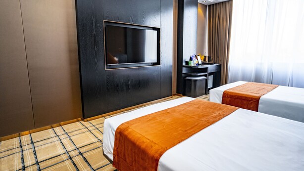 Quarto de hotel com duas camas, uma televisão de tela plana na parede, uma mesinha com abajur e uma janela com cortinas.