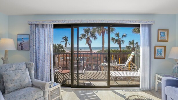 Quarto com uma grande porta de vidro deslizante que dá para uma varanda com vista para palmeiras e o oceano. Há duas cadeiras e uma mesa pequena na varanda.