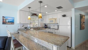Uma cozinha moderna com bancadas de granito, armários brancos e eletrodomésticos de aço inoxidável. Há luminárias pendentes, um micro-ondas, um fogão e uma geladeira.