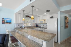 Une cuisine moderne dotée de plans de travail en granit, de meubles blancs et d’appareils électroménagers en acier inoxydable. On y trouve des luminaires suspendus, un four à micro-ondes, une cuisinière et un réfrigérateur.