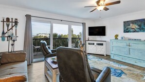 A spacious living room with a large sliding glass door leading to a balcony, a ceiling fan, a flat-screen TV, and a light blue wooden cabinet. - 🌅 Gulf Gate Resort Unit 206 – Updated Bay View Condo with Coastal Charm for Up to 4 Guests (St Pete Beach)