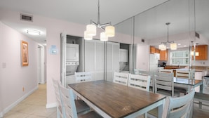 A dining area with a wooden table and white chairs, a kitchen with wooden cabinets, and a mirrored wall reflecting the room. - 🌅 Gulf Gate Resort #508 – Dual Water Views & Coastal Comfort (St Pete Beach)