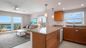 A spacious living area with a large island, a balcony with a view, and a kitchen with wooden cabinets and a window. - 🌅 Gulf Gate Resort #508 – Dual Water Views & Coastal Comfort (St Pete Beach)