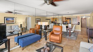 A spacious living room with a large television, comfortable seating, a ceiling fan, and a dining area with a table and chairs. - 🌅 Unit 102 at Gulf Gate – Renovated Luxury with Gulf Views for Up to 6 Guests (St Pete Beach)