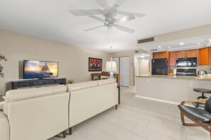 A modern living room with a sofa, a television, a ceiling fan, and a kitchen area with wooden cabinets and appliances.