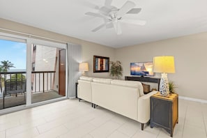 A living room with a sliding glass door leading to a balcony, a ceiling fan, a sofa, a TV, and a lamp.