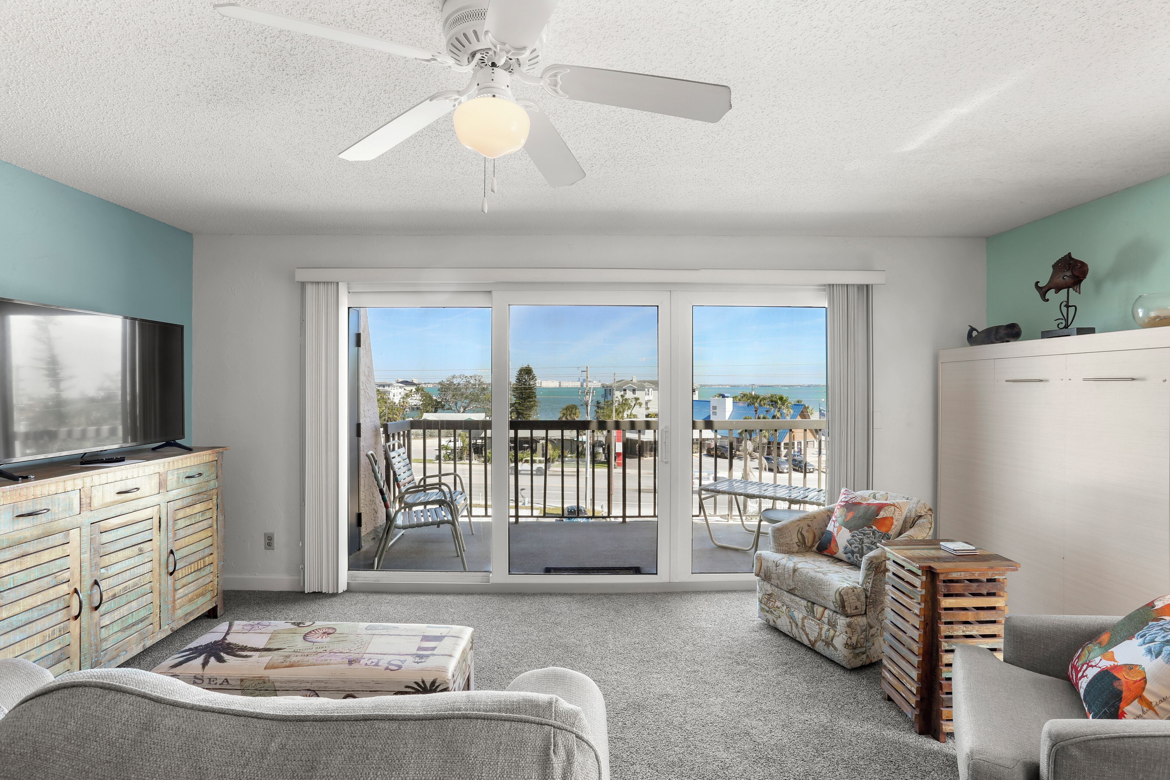 A spacious living room with a large TV, comfortable seating, a ceiling fan, and a balcony with a view.