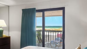 A hotel room with a bed, a nightstand, a lamp, a window with a view of the beach, and a balcony with chairs and a table. - 🌅 Gulf Gate Resort Unit 208 – Dual-Balcony Water View Condo for Up to 4 Guests (St Pete Beach)