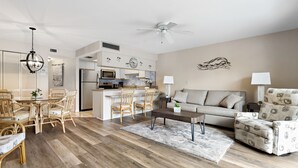 A modern living room with a sofa, armchair, dining table, and chairs. There is a kitchen area with a refrigerator, microwave, and sink. - 🌿 Unit 107 at Gulf Gate – Peaceful Garden View Comfort for Up to 4 Guests (St Pete Beach)