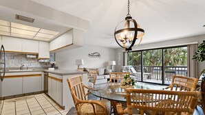 A modern kitchen with white cabinets, a dining area with a glass table and wicker chairs, and a balcony with a view of palm trees. - 🌿 Unit 107 at Gulf Gate – Peaceful Garden View Comfort for Up to 4 Guests (St Pete Beach)