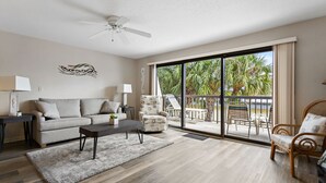 A spacious living room with a sofa, coffee table, and chairs, a ceiling fan, and a large sliding glass door leading to an outdoor area with a pool and palm trees. - 🌿 Unit 107 at Gulf Gate – Peaceful Garden View Comfort for Up to 4 Guests (St Pete Beach)