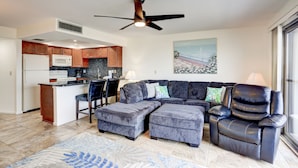 Ein modernes Wohnzimmer mit einem Ecksofa, einem Hocker und einem schwarzen Ledersessel. Es gibt eine Küchenzeile mit Holzschränken und einer Mikrowelle. Ein Deckenventilator ist sichtbar, und ein Gemälde hängt an der Wand.