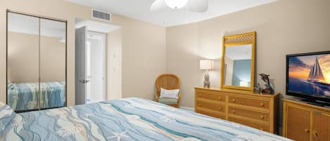 Ein Schlafzimmer mit einem Bett, einem Fernseher, einem Schminktisch, einem Sessel, einem Spiegel und einem Deckenventilator.