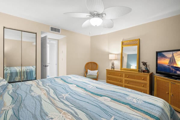 A bedroom with a bed, a television, a dresser, a chair, a mirror, and a ceiling fan.