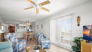 A living room with a ceiling fan, two floral armchairs, a coffee table with a plant, a dining area with a table and chairs, and a kitchen in the background. - !!!Newly Updated, Throughout!!! Gulf Gate 305 | Bay View Family Escape (St Pete Beach)