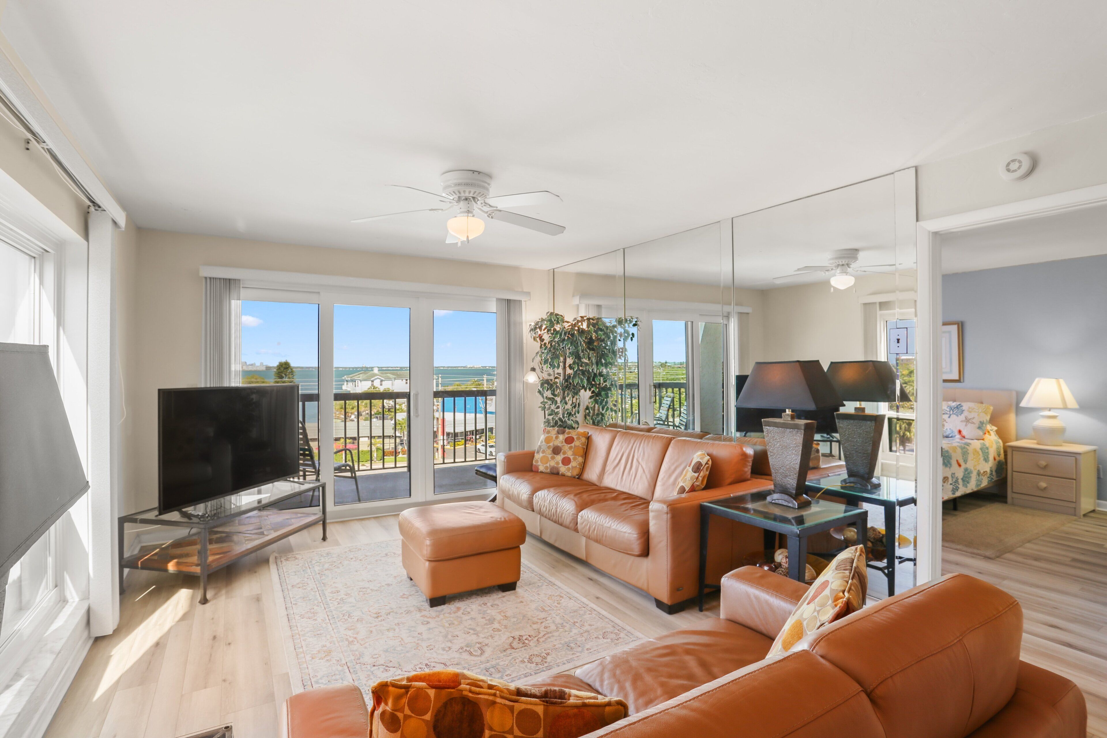 A spacious living room with a large flat-screen TV, leather sofas, a glass coffee table, and a balcony with a view.