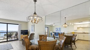 A spacious living area with a glass dining table surrounded by wicker chairs, a balcony with a view, and a kitchen with white cabinets.