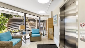 A modern hotel lobby with wicker furniture, a glass partition, and a visible elevator.