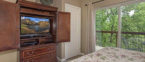 A bedroom with a large bed, a wooden entertainment center with a television, a door, and a balcony with a view of trees.
