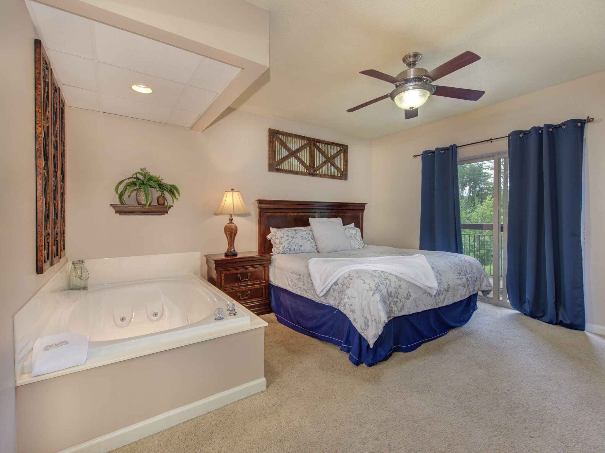 A bedroom with a large bed, a separate bathroom with a bathtub, a ceiling fan, and a view through a window with curtains.