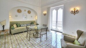 A room with a glass-top dining table, a sofa, and a chair. There is a lamp on a side table, and a balcony with a railing is visible through the window.