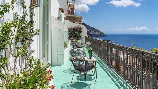 A balcony with outdoor seating, a view of the sea, and a white building with a balcony railing.