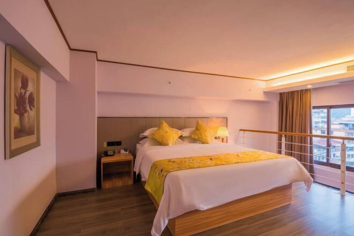 A modern hotel room with a large bed, a nightstand, a window with a view, and a painting on the wall.