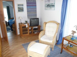 Ein Wohnzimmer mit Fernseher auf Holzständer, ein Korbsessel, ein kleiner Hocker und einem Tisch mit Vase und Rubik's Cube.
