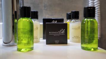 A hotel bathroom with a mirror, a hairdryer, and various toiletries including green bottles and a black box labeled 'Business Life Hotel'.