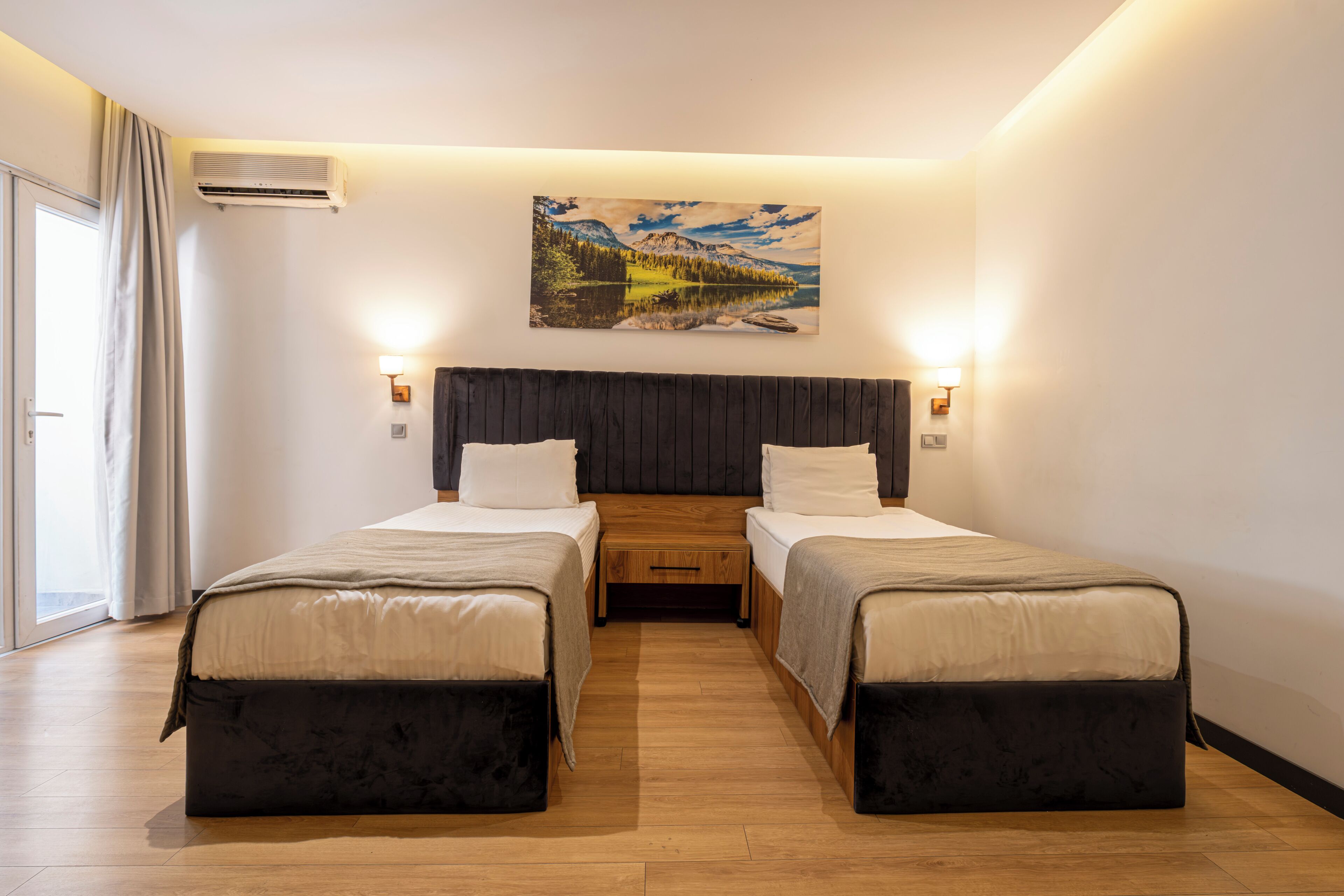 A hotel room with two beds, a wooden floor, a painting on the wall, and a window with curtains.