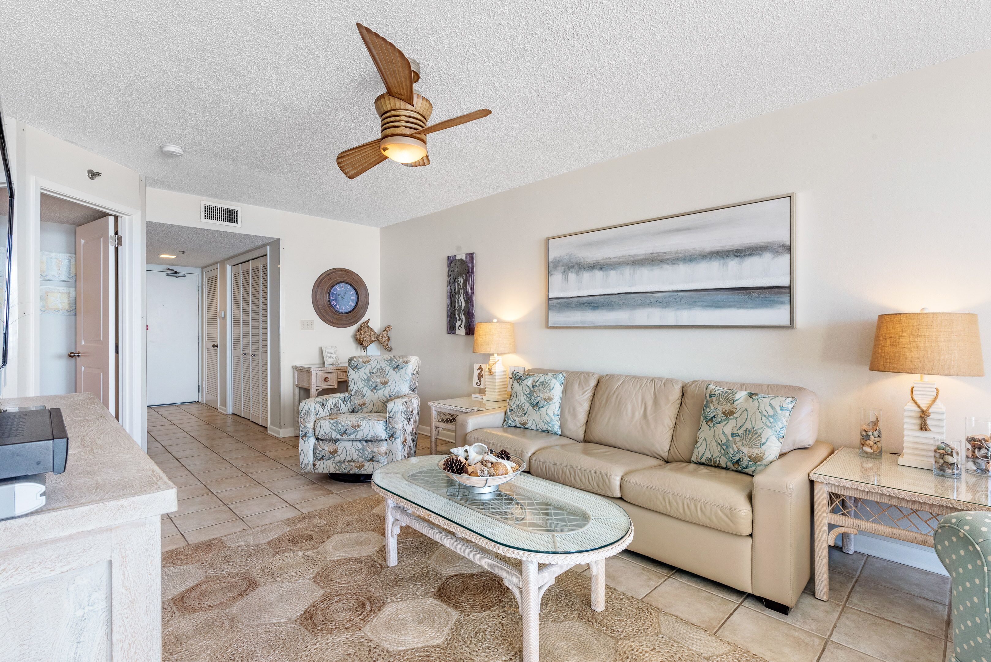 A living room with a beige sofa, a glass coffee table, a ceiling fan, a painting, and a side table with a lamp.