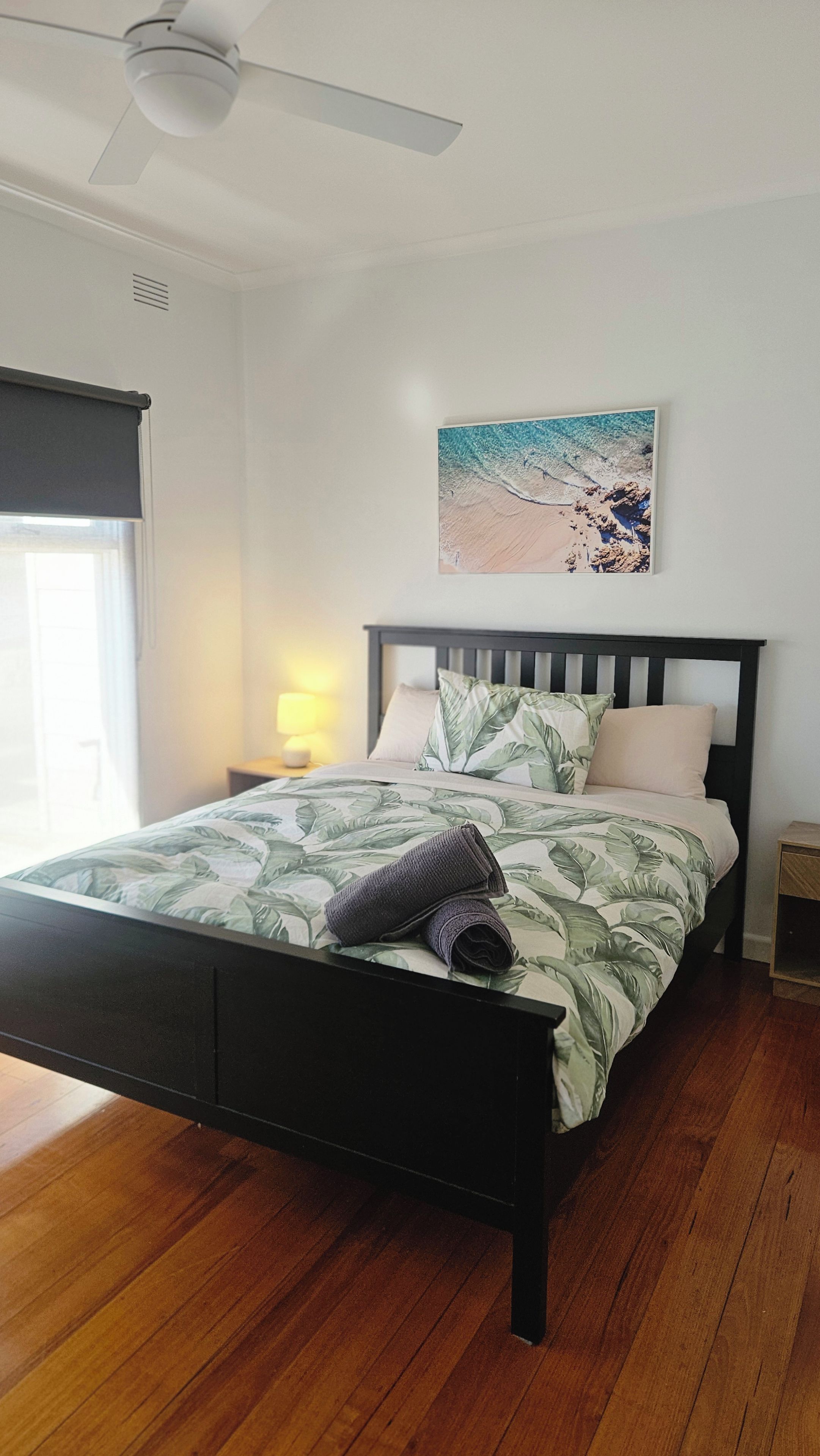Una camera da letto con un letto, una ventola a soffitto, un comodino con una lampada e un quadro a tema spiaggia.