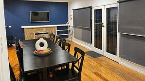 Uma sala de jantar com mesa e cadeiras, lareira, televisão e janelas amplas.