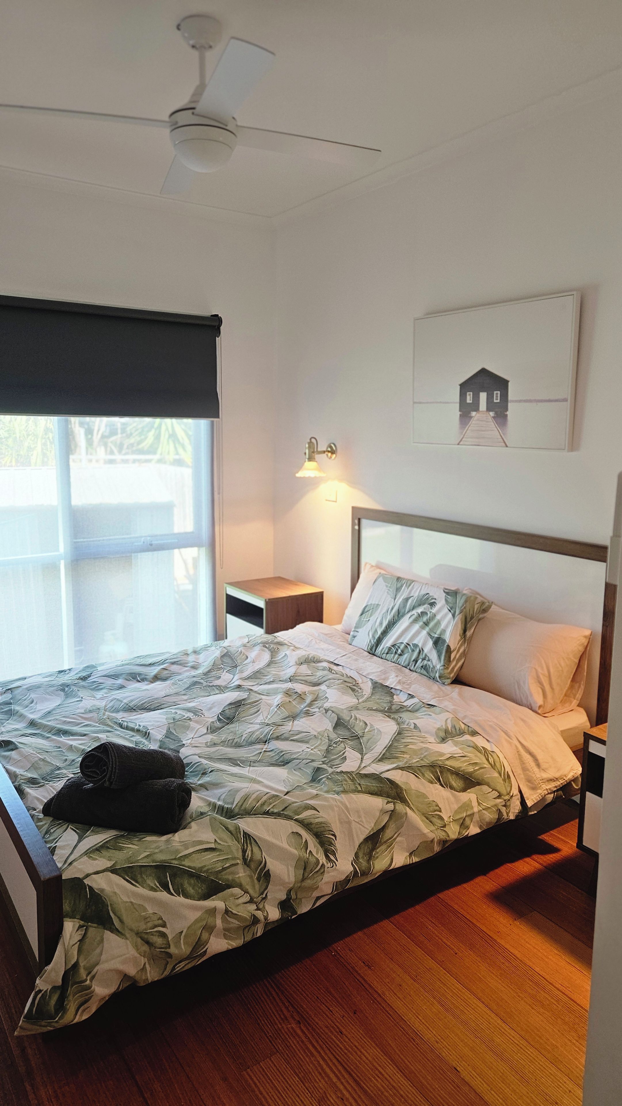 A bedroom with a bed, a nightstand, a wall-mounted lamp, a ceiling fan, and a window with a blind.