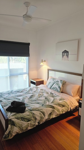 Une chambre à coucher comprenant un lit, une table de chevet, une lampe fixée au mur, un ventilateur de plafond et une fenêtre avec un store.