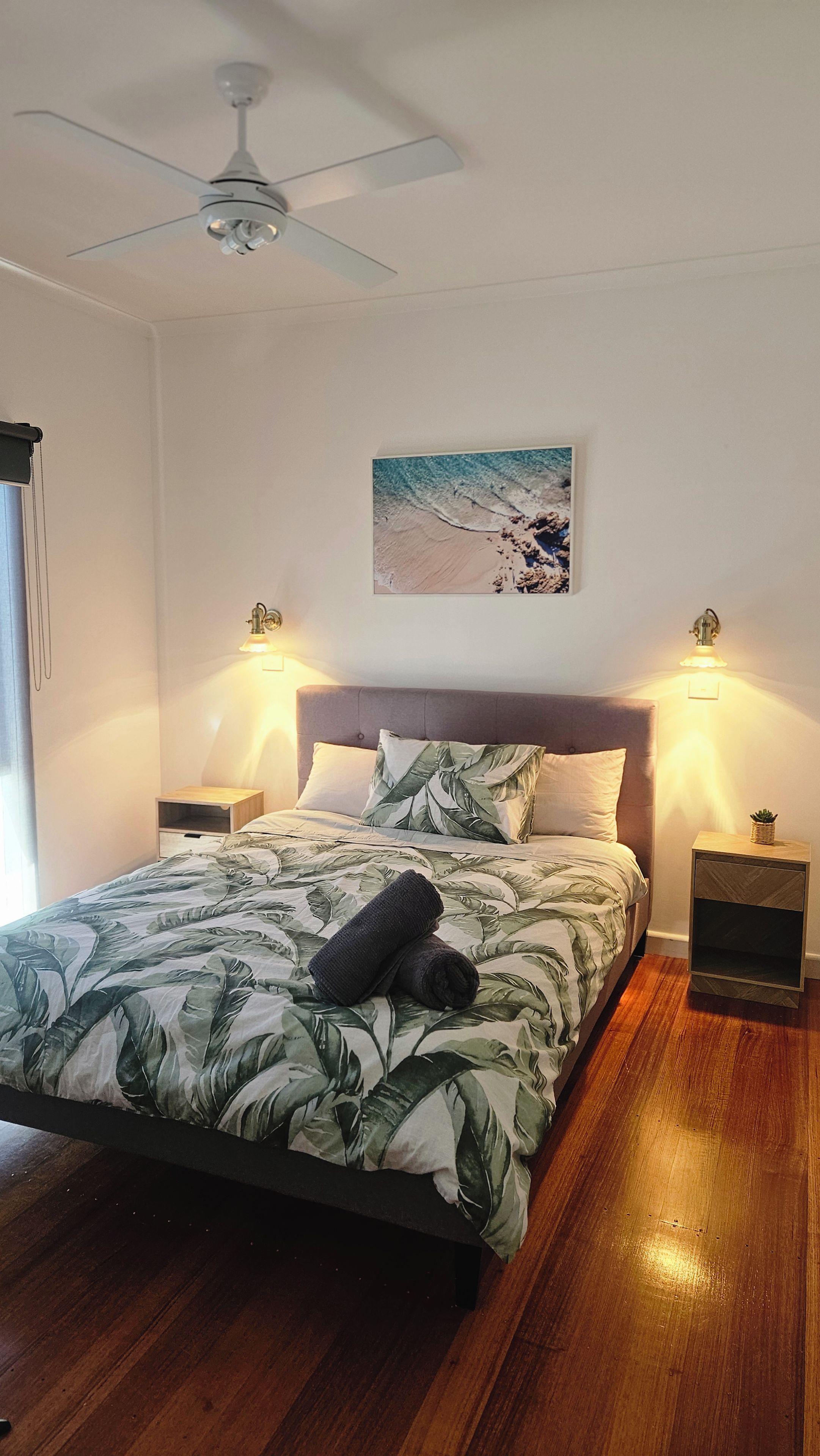 A bedroom with a bed, a ceiling fan, two bedside lamps, a wall-mounted picture, and a wooden floor.