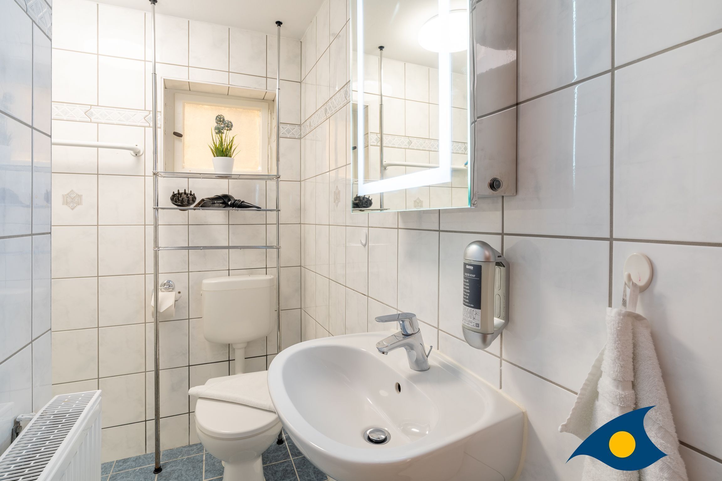 A modern bathroom with a white sink, toilet, and shower. There is a shelf with decorative items and a mirror above the sink.