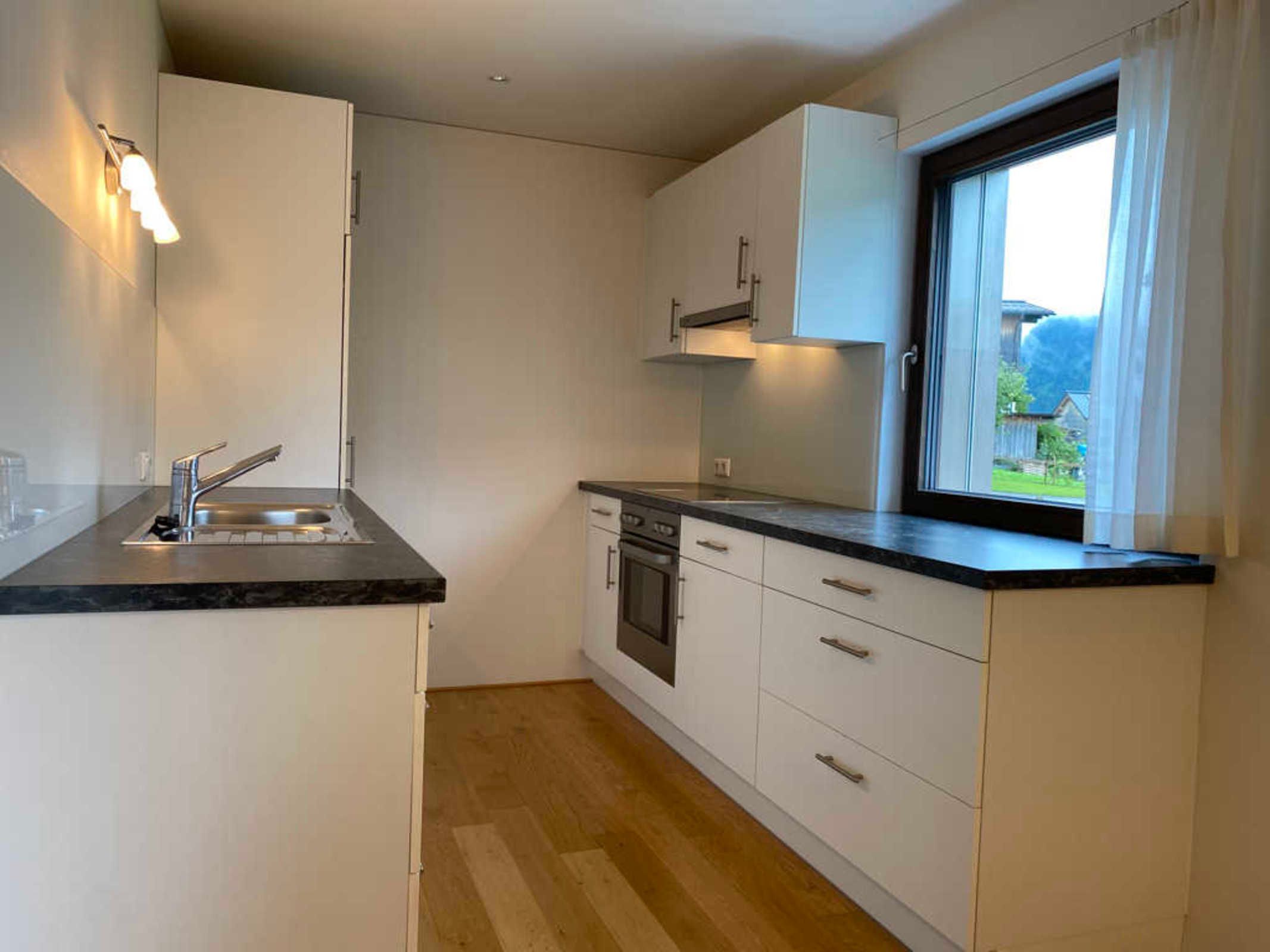 A compact kitchen with white cabinets, a black countertop, a window with a view, and a wooden floor.