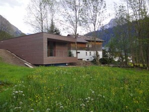 A modern two-story house with a sloped roof, surrounded by a lush green field and mountains in the background.