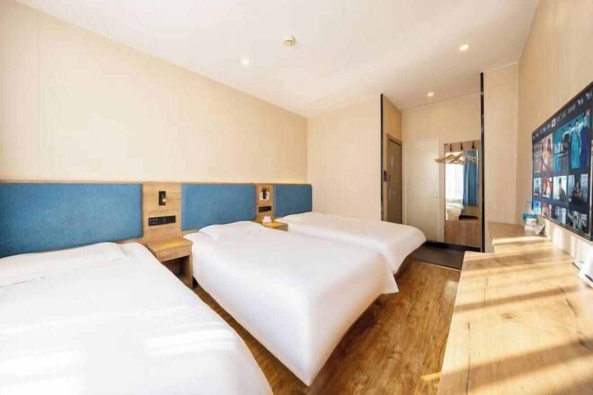 Habitación de hotel con tres camas individuales, suelo de madera, un espejo grande y un televisor montado en la pared.