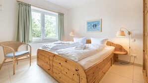 A bedroom with a wooden bed, a wicker chair, a bedside table with a lamp, and a window with curtains. - House half 11 Dune 11 (ID 420) (Sankt Peter-Ording)