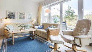 A cozy living room with a sofa, armchair, and coffee table, large windows with a view of greenery, and wall-mounted artwork. - House half 11 Dune 11 (ID 420) (Sankt Peter-Ording)