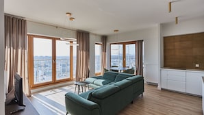 Un salon moderne avec un canapé vert, une petite table et un téléviseur. De grandes fenêtres offrent une vue sur la ville.