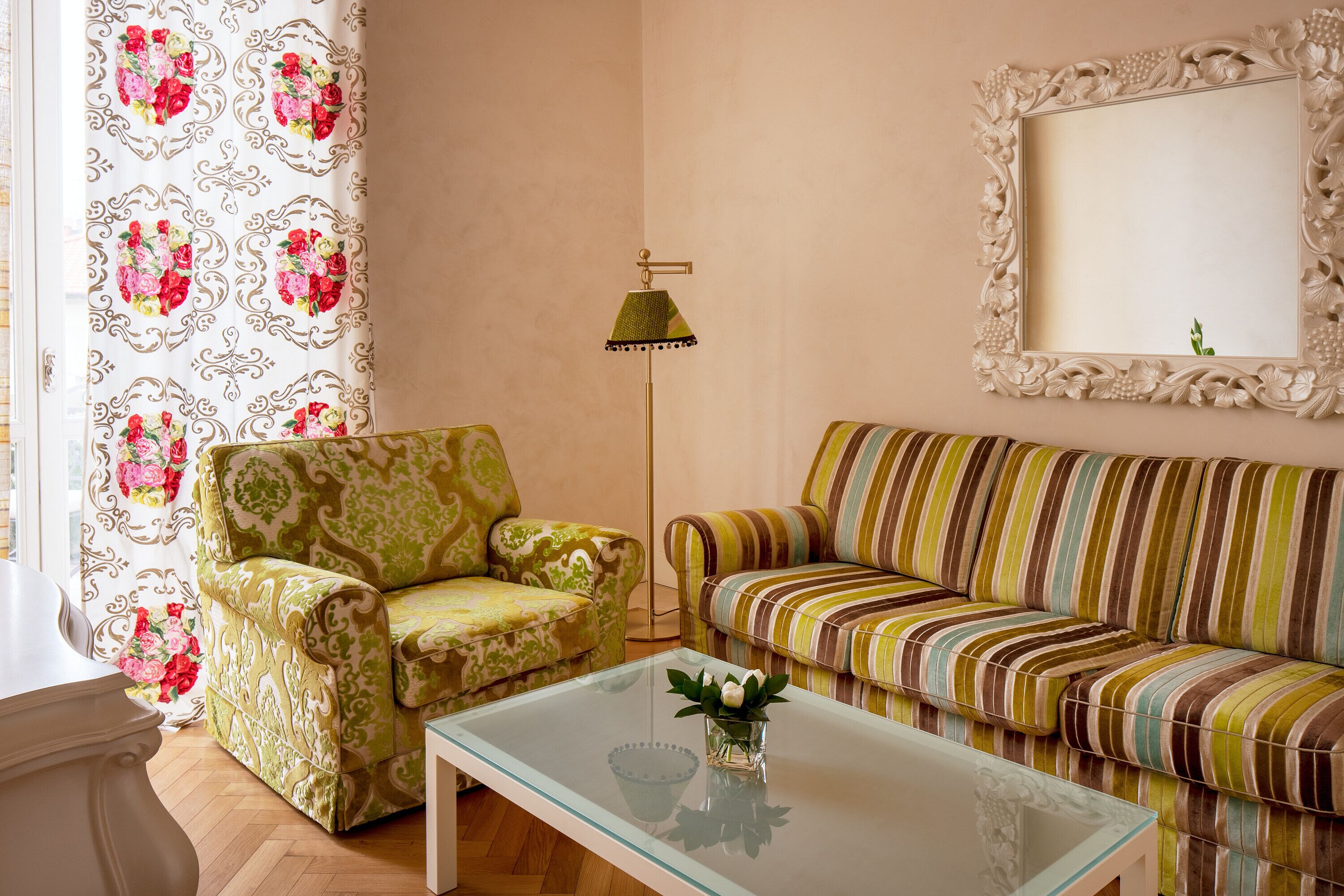Una sala de estar con un sillón estampado, un sofá a rayas, una mesa de centro de vidrio y una lámpara de pie.