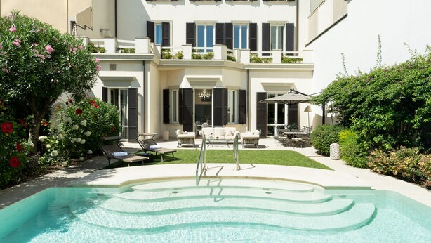 Un edificio de dos plantas con piscina, mobiliario exterior y abundante vegetación.